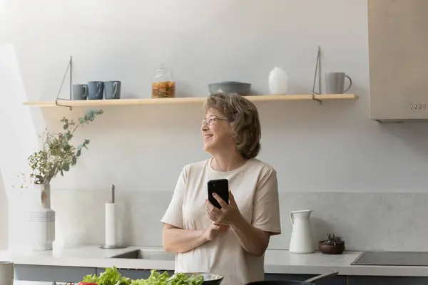 Sonriente soñadora mujer mayor madura utilizando el teléfono celular en la cocina. — Foto de Stock