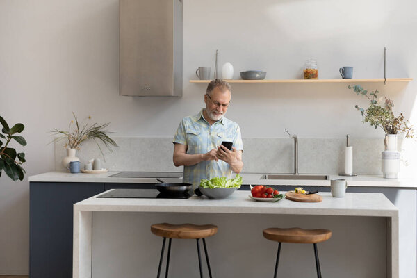 Happy middle aged 60s man using cellphone cooking in kitchen.