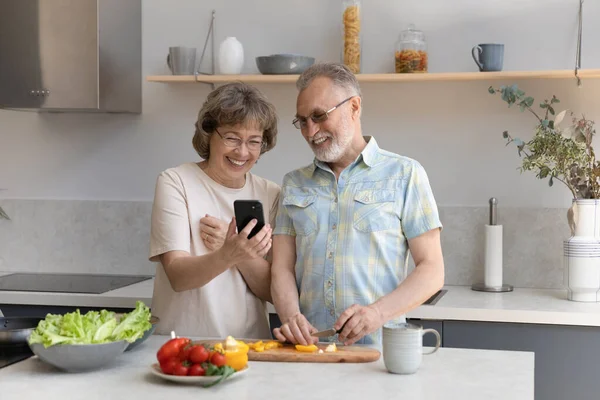 Cheerful old couple using cellphone cooking in kitchen. — Stock Fotó