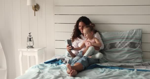 Mom and little daughter rest on bed using smartphone — Vídeo de Stock