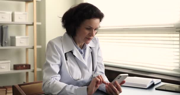 Mature female physician in coat sit at desk holds cellphone — Stockvideo