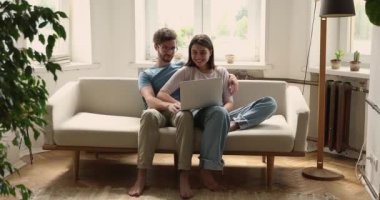 Attractive millennial couple relaxing on cozy sofa with laptop