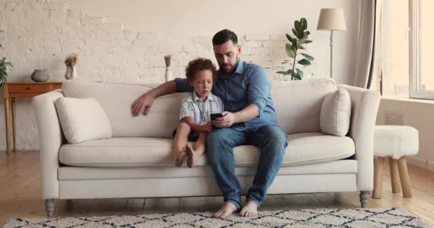 Dad and 5s son sit on couch using smart phone — Αρχείο Βίντεο