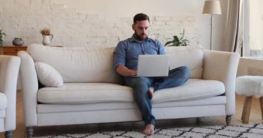 Serious freelancer man sit on sofa work remotely use laptop