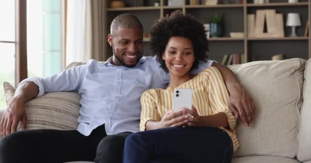 Smiling African American family couple use phone on comfy couch — 图库视频影像