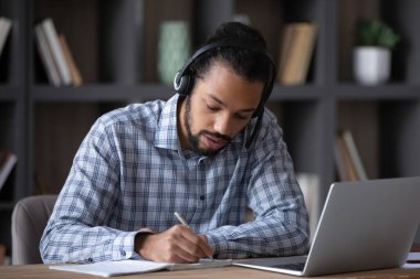 Focused student guy in headphones with mic writing notes