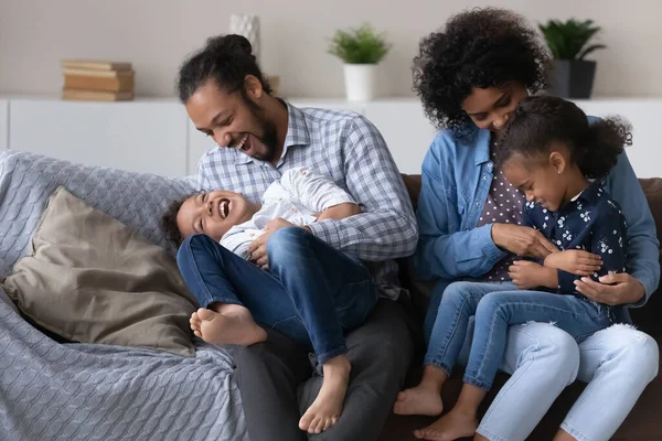 Excited joyful African parents cuddling, tickling kids on couch — ストック写真