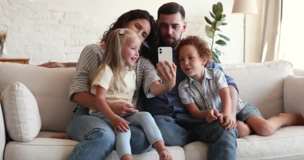 Сім'я з братами і сестрами, які розслабляються на дивані, весело користуються мобільним телефоном — стокове відео