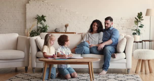 Little kids drawing with pencils, their parents relaxing on sofa — Αρχείο Βίντεο