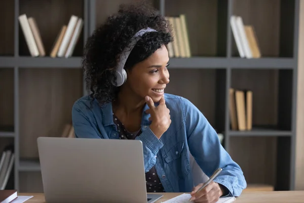 Thoughtful happy African student girl in wireless headphones listening lesson — стоковое фото