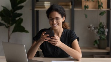 Smiling 30s young businesswoman using smartphone, sitting in office.
