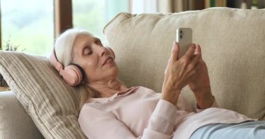 Peaceful older woman wear headphones holds cellphone relax on sofa