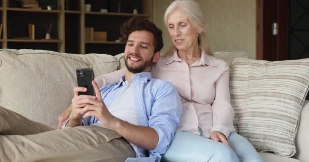 Grownup son older mum relax on sofa using cellphone — ストック動画