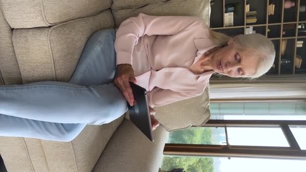Vertical view mature woman sit on sofa using digital tablet — стоковое видео