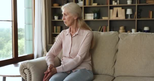 Pensive older female looks into distance sit on sofa indoors — 비디오