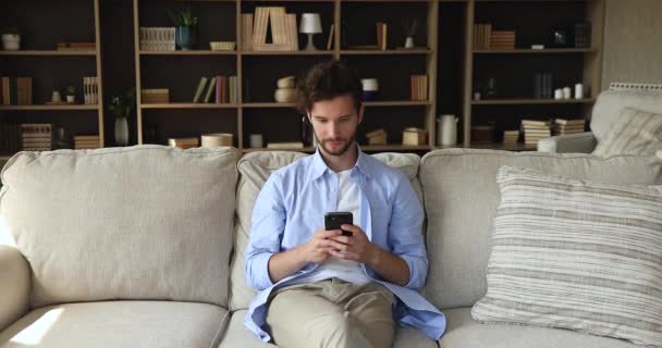 Young guy sit on couch holds cellphone watching online content — ストック動画