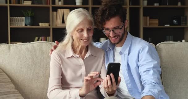 Adult son and older mom sit on sofa using cellphone — Vídeos de Stock