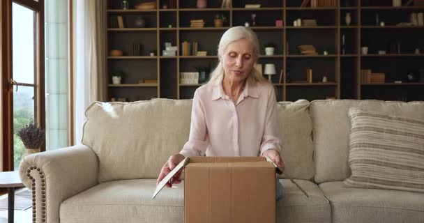 Older woman sit on couch unpack parcel box with books — Video