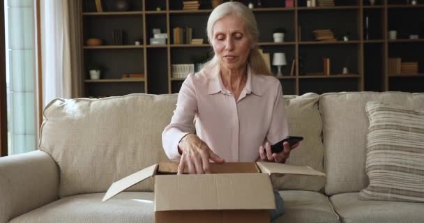 Older woman unpack parcel use cellphone check ordered books online — стоковое видео