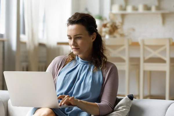 Mujer joven pensativa usando la computadora sentada en el sofá. — Foto de Stock