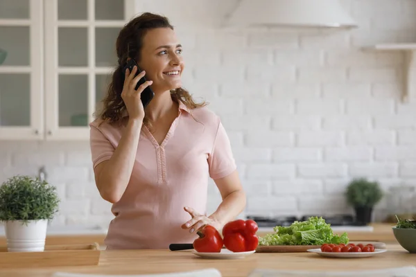 Smiling young woman holding cellphone call conversation, cooking food at home. — Stockfoto
