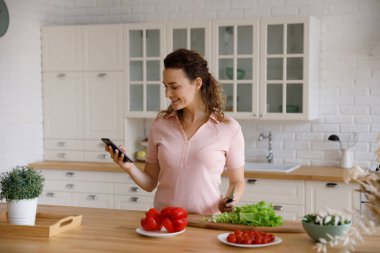 Happy beautiful young woman using cellphone, cooking food in kitchen.