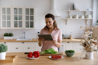 Joyful young woman using digital computer tablet, preparing food at home.