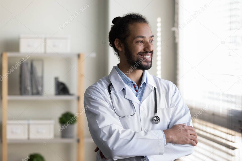 Happy dreamy young African American male doctor standing in office.