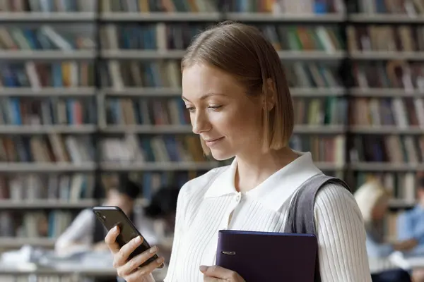 Chica estudiante positiva utilizando la aplicación en el teléfono inteligente en la biblioteca de la universidad —  Fotos de Stock
