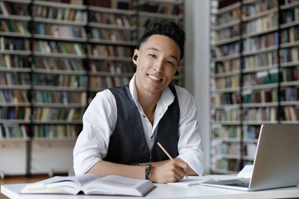 Felice studente asiatico con auricolari wireless studiare in biblioteca headshot — Foto Stock