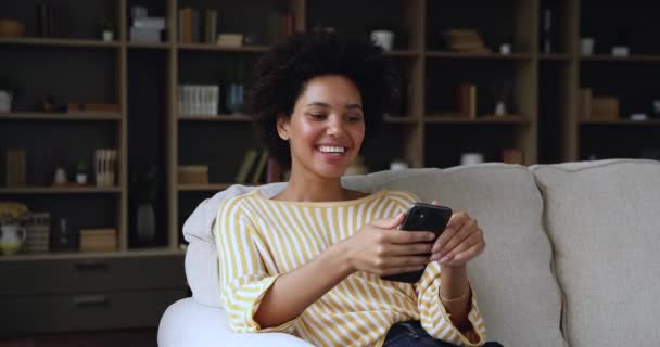 Afrikanerin sitzt auf Sofa und verbringt Freizeit mit Smartphone — Stockvideo