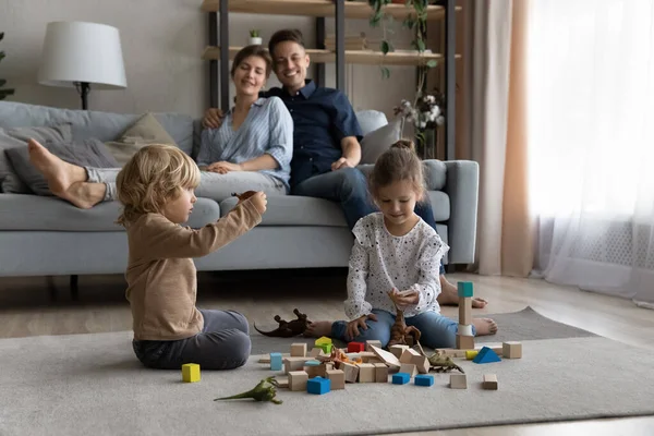 Šťastné malé děti hrají hračky, zatímco rodiče odpočívají na pohovce. — Stock fotografie