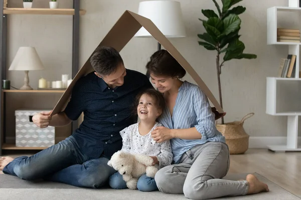 Glad bindning unga föräldrar sitter under kartong tak med dotter. — Stockfoto