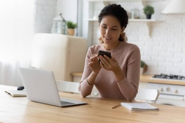 Çekici 30 'lu yaşlarda Latin bir kadın akıllı telefonuyla mutfakta oturuyor.