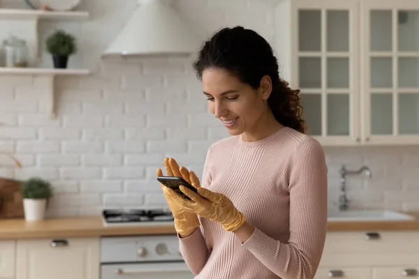 Latina ama de casa en guantes de protección distraída de las tareas domésticas usando smartphone —  Fotos de Stock