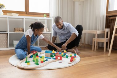 Genç kız ve babası evde küp ve demiryolu oynuyorlar.
