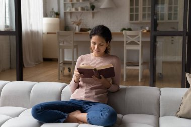 Kitap okuyan genç bir kadın evdeki kanepede bağdaş kurup oturur.