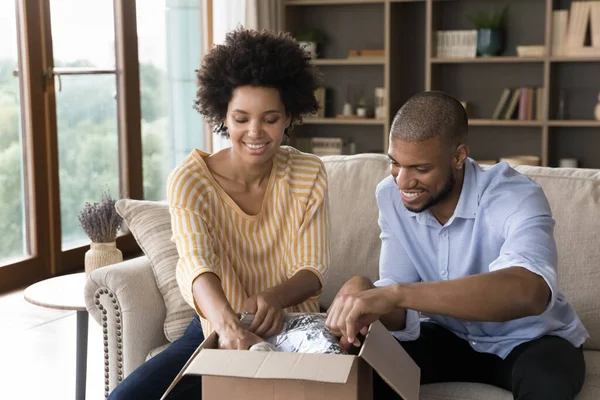 Feliz africano pareja desembalaje paquete sentarse en sofá en casa —  Fotos de Stock