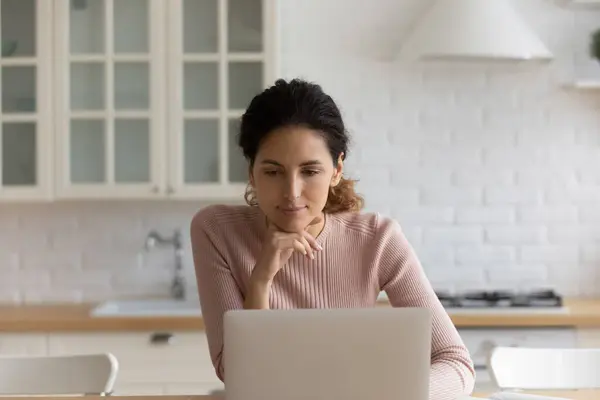 Spaanse vrouw zitten in keuken web surfen met behulp van laptop — Stockfoto