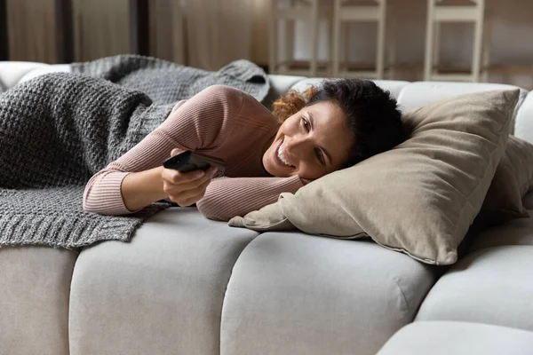 Mujer sonriente acostada en el sofá sostiene canales de interruptor de control remoto — Foto de Stock