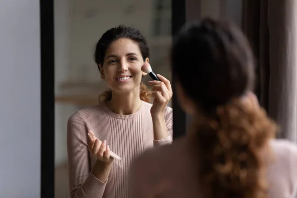 Mujer mira en espejo sostiene cepillo aplicar polvo en los pómulos — Foto de Stock