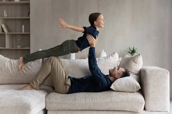 Pai feliz deitado no sofá, levantando filha fingindo voar — Fotografia de Stock