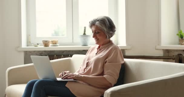 Oudere vrouw zit op de bank met laptop genieten van communicatie online — Stockvideo