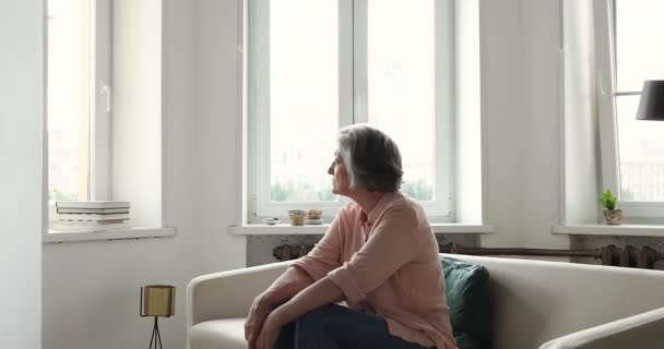 Ernstige oudere vrouw staren naar buiten raam zitten op de bank binnen — Stockvideo