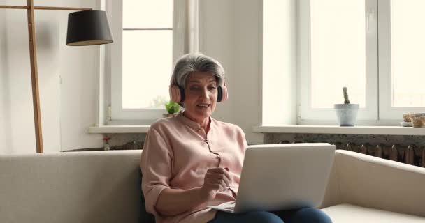 ヘッドフォンの高齢女性は自宅でビデオ会議を使用して話す — ストック動画