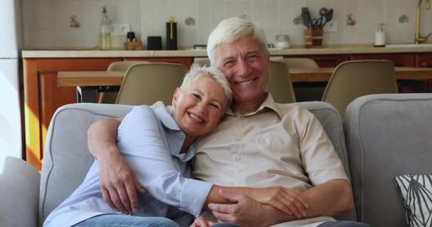 Смеющаяся пожилая пара обнимается расслабляясь на диване, смотрит в камеру — стоковое видео