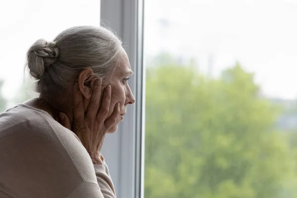 Расстроенная одинокая старшая леди, выглядывающая в окно с отчаянием — стоковое фото