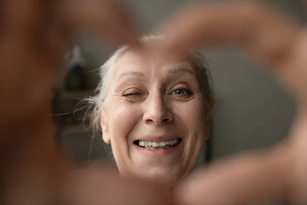 Šťastný senior dáma mrkání na kameru přes ruku prst srdce — Stock fotografie