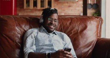 Kulaklıklı gülümseyen Afrikalı adam kanepede telefonla rahatlıyor.