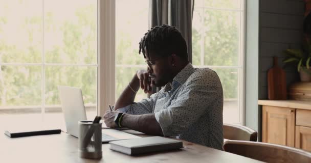 Czarny mężczyzna nauka przez laptop w kuchnia pisać w copybook — Wideo stockowe